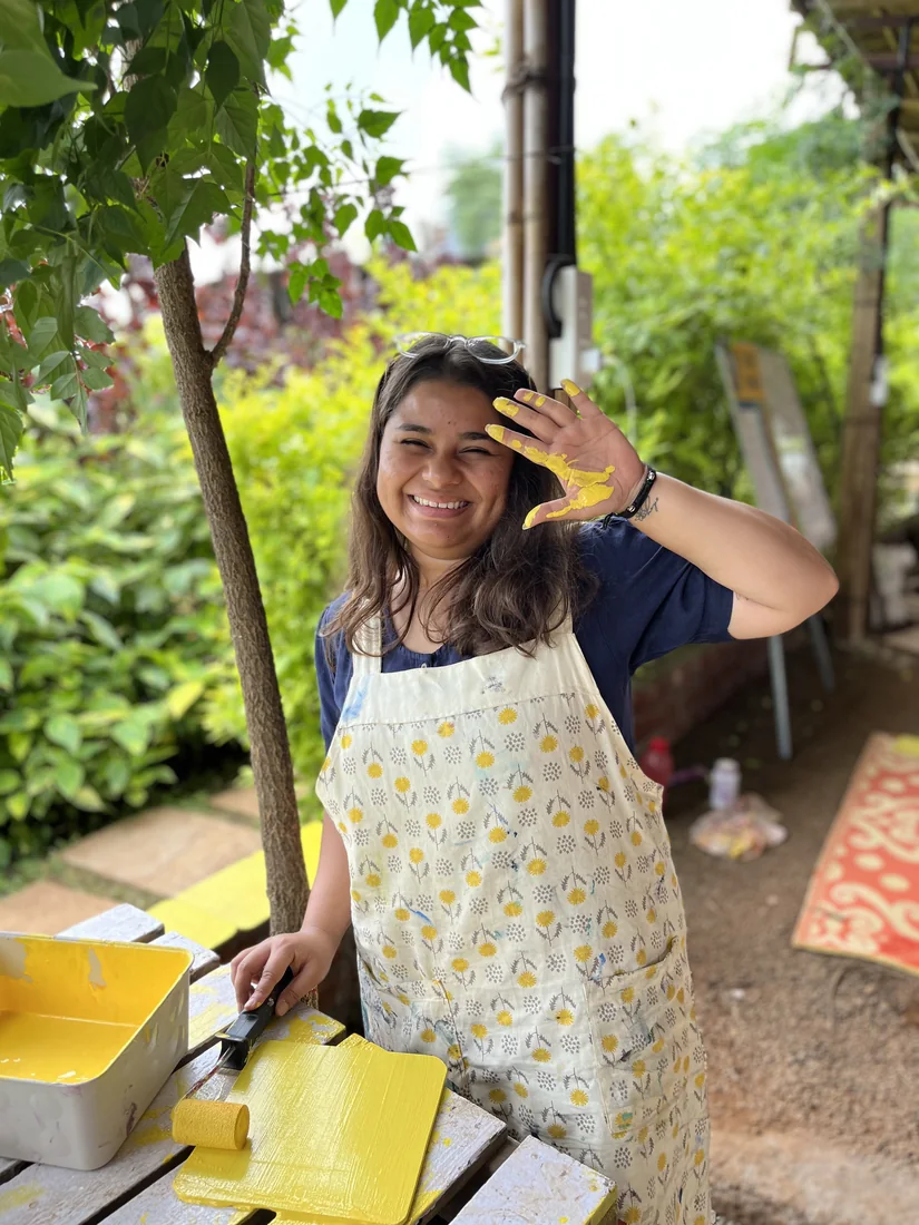 Muskan Mandot laughing, one hand covered in yellow paint, rolling paint onto a wooden board.